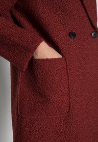 Burgundy texturerad kappa med dubbelknäppt design, utrustad med svarta knappar och en sidficka, som framhäver det mjuka materialet.