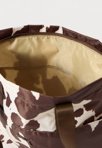 Sac fourre-tout marron et crème avec motif imprimé vache, fermeture éclair, et intérieur beige avec un motif de logo subtil. Tissu durable, forme structurée.