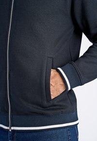 Felpa nera con zip realizzata in tessuto morbido, con polsini e orlo a coste, dotata di una striscia d'accento bianca e una tasca laterale.