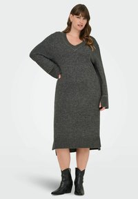 Lång grå stickad klänning med V-ringning, översize ärmar, ribbad botten och dekorativa detaljer längs utrigningen, i kombination med svarta ankelstövlar.