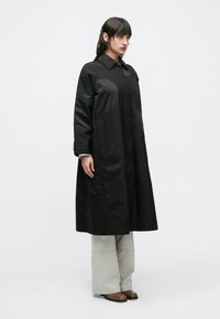 Femeie purtând o haină lungă neagră peste pantaloni largi gri deschis și încălțăminte maro, stând în fața unui fundal simplu și deschis la culoare.