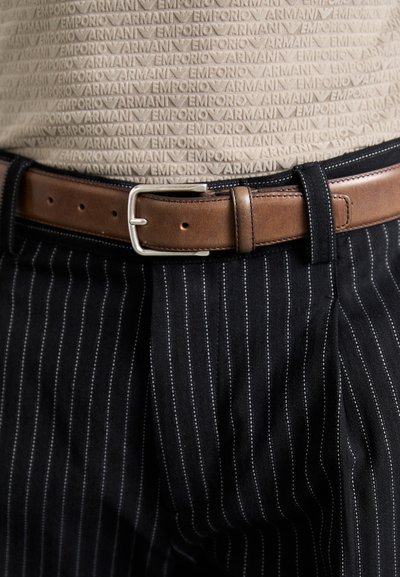 Svart vertikalt stripete bukser med et brunt skinnbelte med en sølvmetallspenne, båret over en teksturert beige topp.