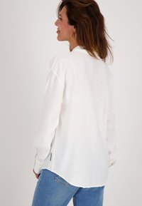 Vrouw met bruin haar draagt gouden oorringen, een witte lange mouwen shirt en blauwe jeans, staande in profiel tegen een effen achtergrond.