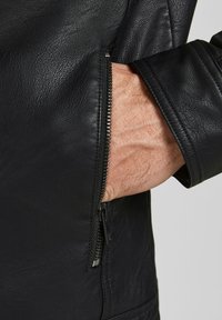 Détail d'une veste en cuir noir montrant un poignet zippé avec une main dans la poche, mettant en avant un cuir texturé et des accents en métal argenté.