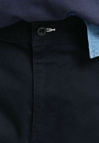 Pantalones azul oscuro con una textura suave, que presentan un botón negro, costuras en contraste y una etiqueta visible con un diseño de espina de pescado.