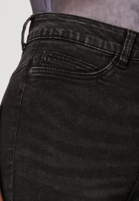 Svarte denimjeans med en falmet tekstur, med høy midje, beltesløyfer og en liten frontlomme; nærbilde av siden.