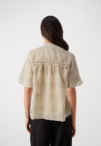 Light beige blouse with short sleeves, featuring floral embroidery, a gathered back, and a lace trim across the yoke. Smooth, sheer fabric.