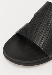 Sandale noire à glissière fabriquée en matériau synthétique texturé, avec une large sangle et une semelle plate. Surface lisse en dessous.