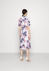 Abito floreale bianco con motivi rosa e blu, maniche corte, vita con cintura e lunghezza midi, abbinato a sandali bianchi con tacco alto.