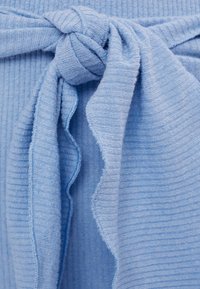 Ljusblått tyg med ribbad textur, med en knuten detalj. Kanterna är något fransade, vilket ger dimension åt designen.