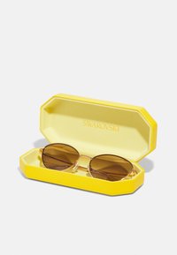 Óculos de sol num estojo octogonal amarelo e texturizado. As lentes são castanhas e a armação é fina em tom de ouro, com elementos de cristal.