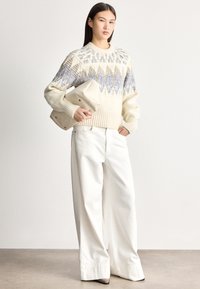 Jovem mulher vestindo um suéter de malha creme com padrão e calças largas brancas, segurando uma carteira grande bege, em pé sobre um chão cinzento.