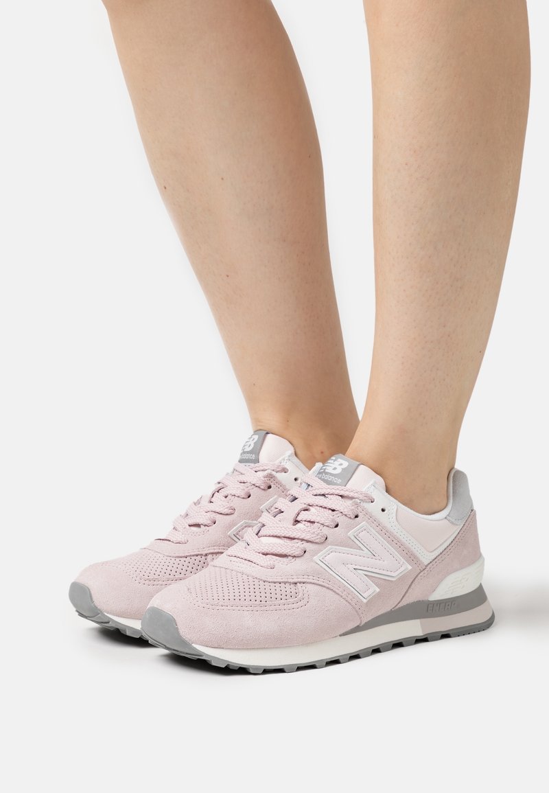 Baskets New Balance rose clair avec des accents gris portées pieds nus, vues de face et de profil sur un fond blanc.