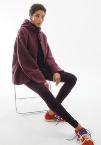 Felpa con cappuccio color granata con zip e coulisse, abbinata a leggings a righe nere e rosso scuro, e sneakers rosse con lacci blu.