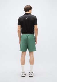 Juoda polo marškinėliai su apykakle ir logotipu nugaroje, derinami su žaliais šortais. Komplektą papildo balti kojinės ir sportbačiai.