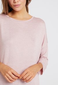 Frau, die ein hellrosa langärmliges Strickoberteil trägt, mit Hängern in den Ohren, die die Hände sanft am Taillenbereich aufeinandergelegt hat, vor einem schlichten Hintergrund.