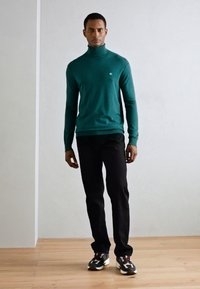 Pull à col roulé vert foncé, design ajusté, associé à un pantalon noir et des baskets. Texture lisse, branding minimal sur la poitrine.