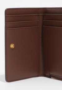 Wnętrze brązowego skórzanego portfela z wieloma slotami na karty, złotym zapięciem na zatrzask oraz wytłoczonym nazwiskiem marki. Gładka tekstura i staranne przeszycia.