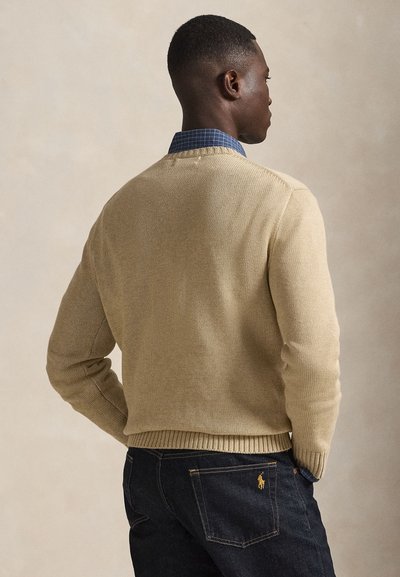 Beige strikkegenser med ribbekant, brukt over en blå rutete skjorte. Denimjeans synlige med en synlig logo på baklommen.