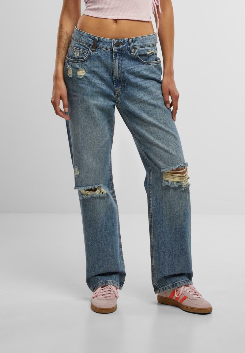 Light blue distressed denim jeans with large rips, wide-leg shape, and five-pocket design. Paired with pink sneakers, featuring a textured surface.