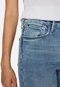 Vaqueros de mezclilla azul claro con un diseño ajustado, que incluyen un bolsillo frontal, trabillas para cinturón y una pequeña etiqueta de marca cerca de la cintura.