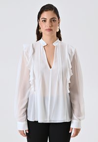 Donna con capelli scuri che indossa una camicetta di chiffon bianca con rouches, scollo a V e maniche lunghe trasparenti, abbinata a pantaloni neri su uno sfondo semplice.