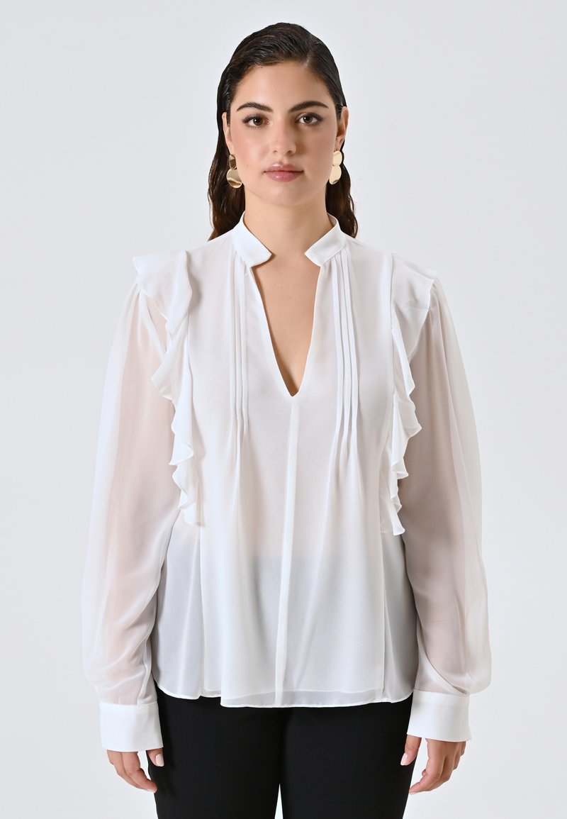 Donna con capelli scuri che indossa una camicetta di chiffon bianca con rouches, scollo a V e maniche lunghe trasparenti, abbinata a pantaloni neri su uno sfondo semplice.