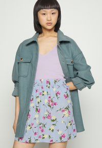 Casaco de tamanho grande em teal com dois bolsos frontais, sobre uma blusa de alças lilás com nervuras, combinado com uma saia estampada em flor azul clara.