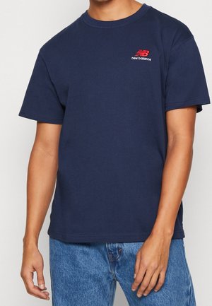 Man draagt een donkerblauw New Balance T-shirt met rood logo op de borst en lichtblauwe spijkerbroek, getoond van schouders tot heupen.