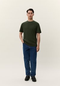 Tmavě zelené tričko s kulatým výstřihem z bavlny, krátké rukávy, doplněné modrými riflemi s rovnými nohavicemi a černými teniskami, stojící na neutrálním pozadí.