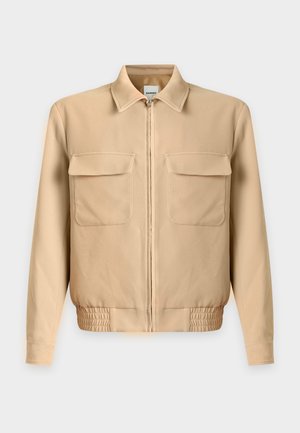 Mann trägt eine beige Jacke mit Reißverschluss und Vorderentaschen, dunkelblaue weite Hose und dunkelbraune Schuhe, steht vor einem schlichten weißen Hintergrund.