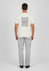 Vit t-shirt med svart mönster på baksidan, med texten "KARL LAGERFELD." Matchas med ljusgrå byxor, enfärgad och med slät textur.