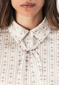 Blouse créme avec un motif brun clair de petits parapluies. Comprend un col rabattu et un détail de nœud noué au niveau du cou.