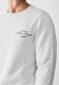 Sudadera gris con cuello redondo acanalado. Presenta texto bordado en negro "COTE D'AZUR Palm Royale" en el lado izquierdo del pecho, con una textura de tela suave.