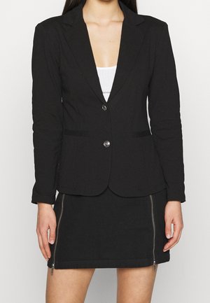 Vrouw draagt een nauwsluitende zwarte blazer met twee knopen over een wit topje, gecombineerd met een zwarte minirok met twee verticale ritsen aan de voorkant.