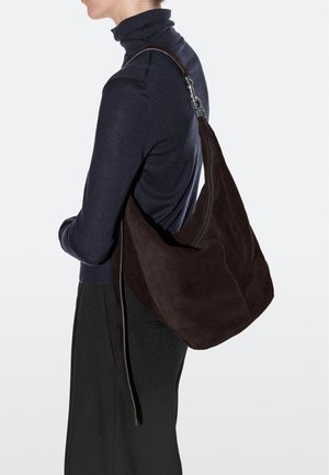 Person trägt schwarzen Rollkragenpullover und Hose und trägt eine große dunkelbraune Wildleder-Schultertasche mit silberner Schnalle am Riemen.