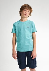 Krátký rukáv mátově zeleného trička s kulatým výstřihem a jemnou texturou, s malým červeným textem na hrudi. V kombinaci s námořnickými cargo šortkami.