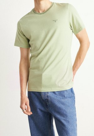 Mężczyzna w jasnooliwkowej koszulce Barbour z krótkim rękawem i niebieskich dżinsach, stojący z jedną ręką w kieszeni, a drugą wzdłuż ciała.