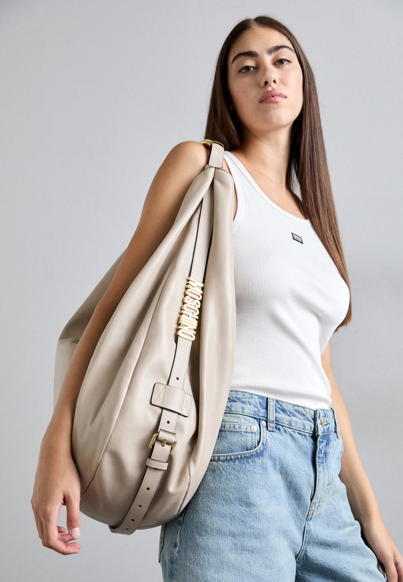 Jauna moteris balta marškinėliais ir mėlynomis džinsomis neša didelę smėlio spalvos Moschino petinę rankinę su auksiniu logotipu ant dirželio.