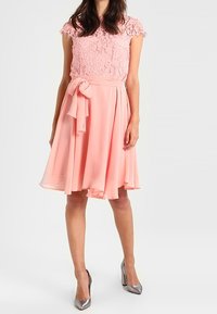 Světle růžové šaty s krajkovým topem, krátkými rukávy a splývavou, délky po kolena, šifonovou sukní. Obsahují látkový pás uvázaný v pase.