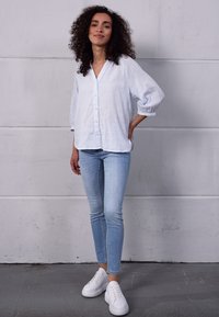 Camisa de botão azul claro com riscas, com mangas relaxadas, combinada com jeans skinny azul claro e ténis brancos. Texturas de tecido suaves.