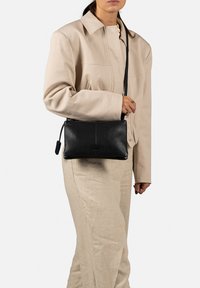 Borsa a tracolla in pelle nera con chiusura a zip, caratterizzata da una texture liscia e una forma rettangolare. La tracolla è fissata su entrambi i lati.