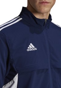 Marineblauwe sportjacket met een ritssluiting aan de voorkant, voorzien van witte strepen op de schouders en een geborduurd Adidas-logo op de linkerkant van de borst.