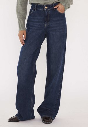 Persona che indossa jeans blu scuro a gamba larga, con una mano in tasca e scarpe marroni visibili sotto i jeans.