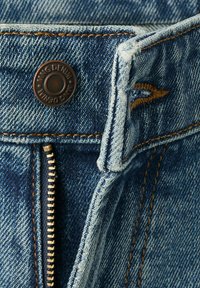 Gros plan sur un jean en denim bleu montrant une fermeture éclair partiellement ouverte, un bouton en métal marqué "MNG DENIM,", des coutures orange et une boucle de ceinture.