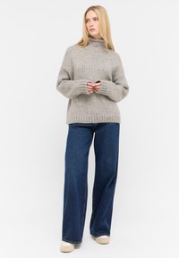 Grauer Strickpullover mit hohem Kragen, lockerer Passform und geripptem Saum, kombiniert mit weit geschnittenen blauen Jeans und Freizeit Schuhen.