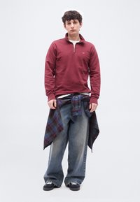 Νεαρός άνδρας που φοράει μπορντό πουλόβερ με φερμουάρ, φαρδιά μπλε τζιν και ένα καρό πουκάμισο δεμένο γύρω από τη μέση του, στέκεται μπροστά σε μια απλή φόντο.