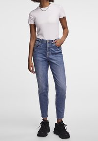 Nainen, jolla on päällään valkoinen lyhythihainen ribbipaita, korkeavyötäröiset siniset farkut, mustat paksupohjaiset lenkkarit ja hopeinen ketjun kaulakoru.