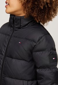 Schwarze Steppjacke mit hohem Kragen und einem Frontreißverschluss. Auffälliges Logoetikett am linken Ärmel, glatte Textur mit gepolsterten Bereichen.