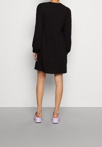 Svart, texturerad klänning med långa ärmar och åtsittande midja; kombinerad med ljuslila klacksandaler. Enkelt, elegant mönster med minimala detaljer.